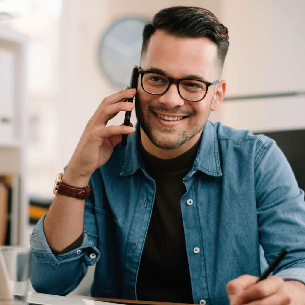 A man in glasses smiling while talking on the phone, seated at a desk with a pen in hand